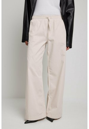 Kvinde iført beige bukser med vidde og elastisk talje, hvid top, sort læderjakke og spidse sorte sko.