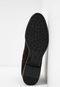 Semelle d'une chaussure en daim marron foncé avec motif de bande de roulement circulaire et talon texturé, présentée debout contre un fond blanc en angle.