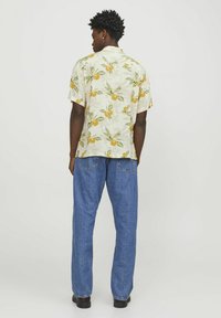 Camisa de manga corta con una tela de color crema claro que presenta un vibrante patrón de hojas en naranja y verde, combinada con unos vaqueros de mezclilla azul sueltos.