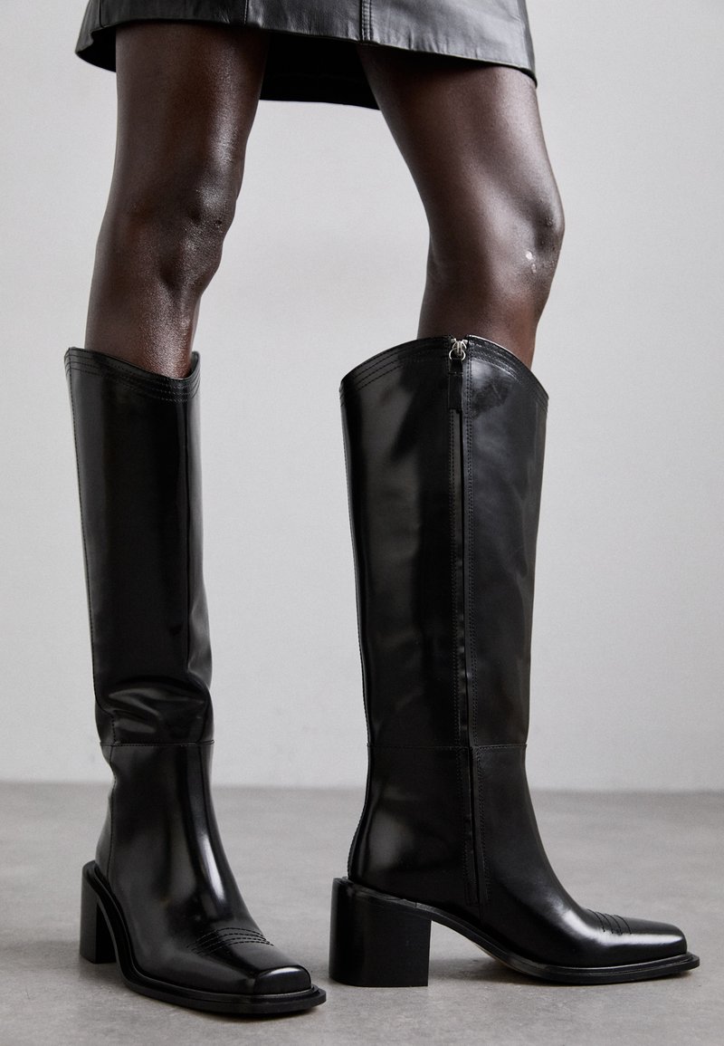 Black knee-high boots with a shiny leather finish, square toe, block heel, and side zipper detail, featuring minimal stitching and a sleek design.
