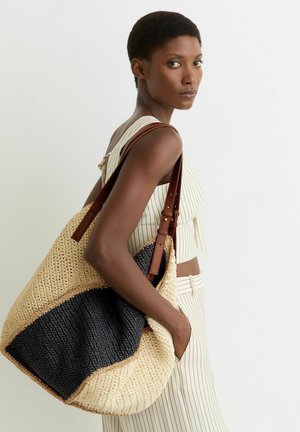 Žena ve smetanovém oblečení s proužky, nesoucí velkou béžovo-černou tkanou tašku s hnědými koženými popruhy přes rameno.