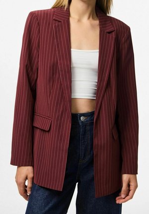 Femme portant un blazer bordeaux à fines rayures sur un crop top blanc et un jean bleu foncé taille haute, debout devant un fond uni.