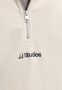 Kreemivärvi villane kangas, millel on osaliselt lahti tõmmatud hõbedane tõmblukk ja "JJ Studios" musta värviga brodeeritud tõmbluku all.