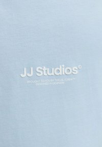 Világoskék szövet fehér felirattal: "JJ Studios", "A JJ Crew™ jóvoltából," és "Dániában tervezve."