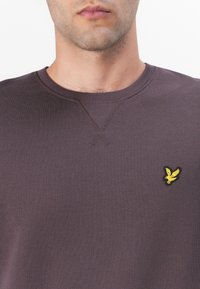 Mørk lilla genser med ribbet rund hals, med en liten brodert gul fuglelogo på venstre bryst, strukturert stoff.