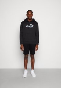 Zwarte hoodie met een voorzak en kleurrijke "silverTab"-tekst, gecombineerd met zwarte shorts en witte sneakers, staande tegen een lichte achtergrond.