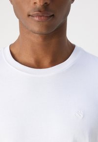 White cotton t-shirt with a round neckline and short sleeves, features a subtle embroidered logo on the chest. Smooth texture.