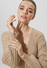 Pull en maille côtelée beige avec un décolleté en V profond ; il a des manches longues avec des nervures texturées. Les accessoires comprennent un collier en chaîne doré et des bagues.