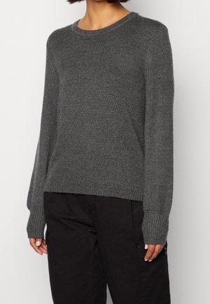 Personne portant un pull en tricot gris foncé à manches longues et un pantalon noir, debout devant un fond clair uni.