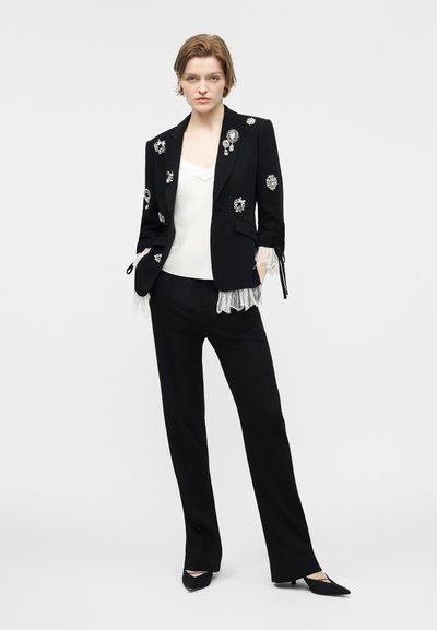 Γυναίκα ντυμένη με μαύρο κοστούμι και διακοσμημένο σακάκι, λευκό πουκάμισο, μαύρα μυτερά παπούτσια, στέκεται με τα χέρια στις τσέπες σε λευκό φόντο.