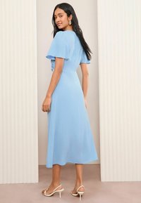 Robe midi bleu clair avec de courtes manches à volants, un corsage ajusté et une jupe fluide. Portée avec des sandales blanches à lanières. Texture lisse.