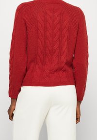 Pull en laine rouge à mailles texturées porté par une personne à la peau foncée avec un pantalon blanc, vu de dos.