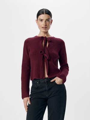 Bordeaux strikket cardigan med cropped længde, prydet med båndlukning ved halsen, parret med sorte jeans. Glat tekstur, tætsiddende design.
