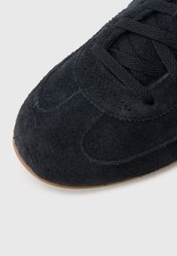 Αθλητικό παπούτσι από μαύρο σουέντ με στρογγυλεμένη μύτη, επίπεδα κορδόνια και υφή στην επιφάνεια· διαθέτει τακούνι από καφέ καουτσούκ στο κάτω μέρος.