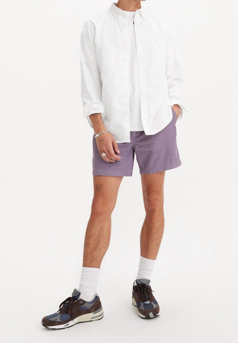 Chemise blanche légère à boutons, portée sur un t-shirt blanc, avec un short violet. Chaussures de sport en matériaux mixtes avec des touches de bleu, marron et gris.