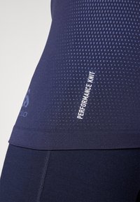 Marineblå prestasjonsstrikket topp med teksturert mønster, elastisk hem og hvit "PERFORMANCE KNIT"-etikett for merkeidentifikasjon.