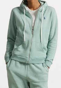 Mintgrön zip-up hoodie i mjukt tyg, med frontdragkedja, vita snörningar och en liten broderad logotyp på bröstet.