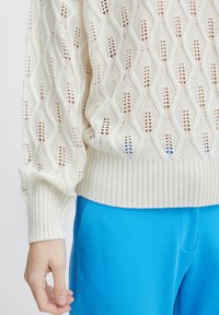 Weißer gestrickter Pullover mit einem strukturierten, offenen Maschenmuster und gerippten Bündchen, kombiniert mit lebhaft blauen Hosen.