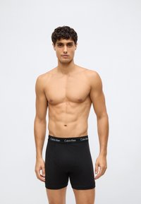 Homem com cabelo curto e encaracolado, sem camisa, a usar boxers pretos da Calvin Klein, em pé contra um fundo claro e liso.