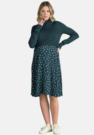 Vrouw in een donkergroene cropped coltrui, donkergroene midi rok met stippen en witte sneakers, staand met één hand op de heup.