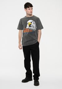 Gray graphic t-shirt featuring a bold eagle design, complemented by the text "Chris Stapleton" in orange. Worn with black pants and boots.