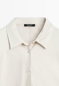 Camicia button-up color crema chiaro con colletto classico, tessuto liscio e tre bottoni. Etichetta con la scritta "Massimo Dutti, MADE IN CHINA."