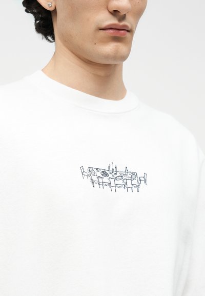 Κοντινό πλάνο ενός ατόμου που φοράει λευκό πουκάμισο με ένα μικρό μαύρο σχέδιο γραμμής ενός τραπεζιού φαγητού με πιάτα, κεριά και καρέκλες.