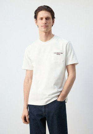 Jeune homme aux cheveux foncés portant un T-shirt blanc avec le texte « Last Check-Out, Please » et un jean bleu foncé, debout devant un fond uni.