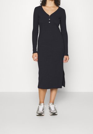 Mujer con un vestido midi negro de manga larga, abotonado, con una abertura lateral y zapatillas deportivas plateadas y blancas sobre un fondo liso.