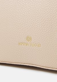 Beige tekstureret syntetisk lædertaske med et gyldent "ANNA FIELD" logo. Blød overflade med et subtile kornmønster og syede kanter.