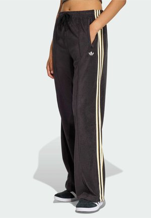Person iført sorte Adidas træningsbukser med cremefarvede sidelister og sorte sneakers, stående med den ene hånd i lommen.