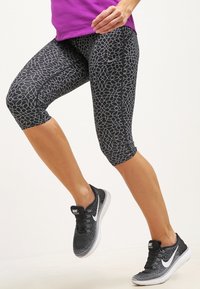 Leggings capri negros con un patrón geométrico blanco, combinados con zapatillas deportivas en blanco y negro. La textura parece suave y ajustada.