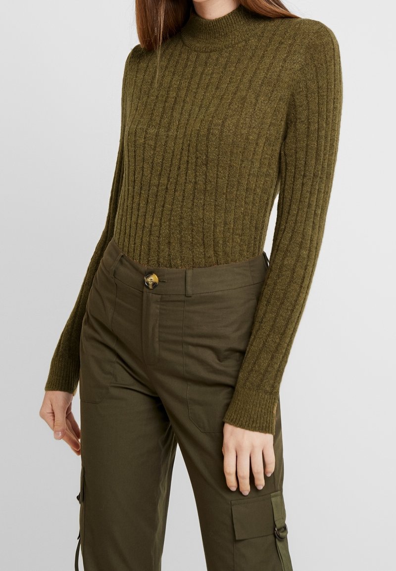 Femme portant un pull en maille côtelée vert olive et un pantalon cargo assorti avec des détails de boutons et de poches, debout devant un fond uni.
