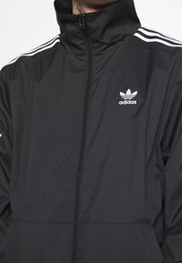 Muž v černé bundě Adidas na zip s vysokým límcem a bílými třemi pruhy na ramenou a logem na hrudi.