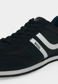 Baskets décontractées noir et blanc avec lacets noirs, empeigne texturée, bande blanche et logo "BOSS" sur le côté.