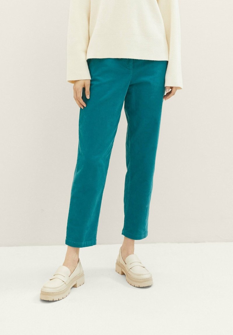 Personne portant un pantalon turquoise à la cheville, un pull oversized crème et des mocassins beiges épais, se tenant sur un sol et un fond clairs.
