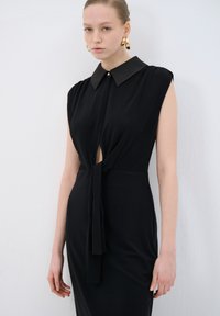 Robe noire sans manches avec un col, présentant un décolleté en keyhole à l'avant et un détail de nœud à la taille. Tissu lisse et ajustement soigné. Accent de bouton doré.