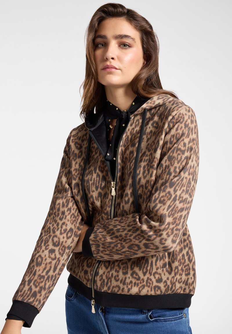 Giovane donna con capelli castani ondulati, che indossa una felpa con zip a stampa leopardata marrone e jeans blu, in posa con le braccia incrociate.