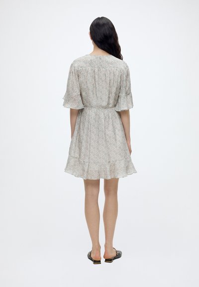 Femme aux longs cheveux foncés portant une robe légère fleurie au genou avec des manches et un ourlet à volants, debout devant un fond blanc uni.