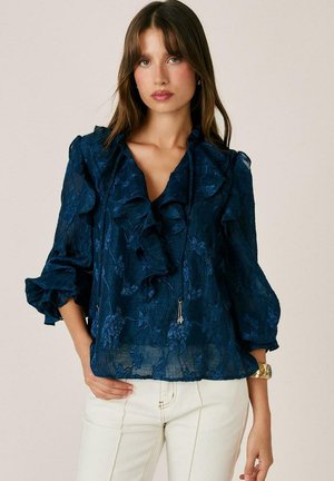 Mujer que lleva una blusa azul oscuro transparente con estampados florales y detalles fruncidos, combinada con pantalones de color claro y un reloj de pulsera dorado.