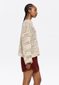 Strickpullover in Off-White mit strukturiertem, offenen Maschenmuster. Abgeschlossen mit einem Rundhalsausschnitt und lockerer Passform, kombiniert mit bordeauxfarbenen Shorts.