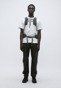 Πράσινο αδιάβροχο γιλέκο πεζοπορίας με ρυθμιζόμενους ιμάντες, φορεμένο πάνω από ένα λευκό πουκάμισο. Λαδί παντελόνια και μαύρα παπούτσια συμπληρώνουν την εμφάνιση. Μινιμαλιστικός σχεδιασμός.