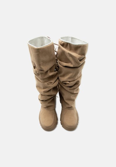 Ψηλές μπεζ σουεντ μπότες με μαλακούς, χαλαρούς κορμούς και ανθεκτική σόλα, βλέποντας από πάνω σε απλό λευκό φόντο.
