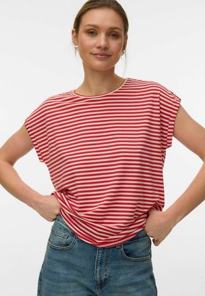 Vrouw met licht haar draagt een rood-wit gestreept T-shirt met korte mouwen en blauwe spijkerbroek, handen op de heupen tegen een effen achtergrond.