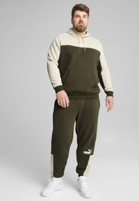 Conjunto de sudadera y pantalones en verde oliva con acentos en crema, que presenta un bolsillo frontal y un logo. La tela parece suave y tiene un corte relajado.