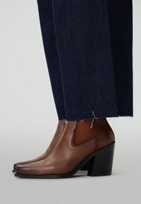Bottines en cuir marron avec un bout carré, des panneaux latéraux élastiques et un talon épais noir. Associées à un pantalon en denim foncé avec un ourlet effiloché.