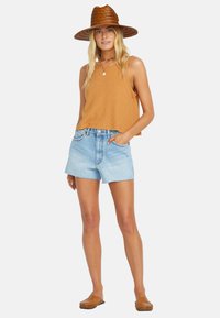 Hnedý rebrovaný crop top, svetlomodré džínsové šortky s roztrhnutým lemom, hnedé šľapky a široký slamenný klobúk. Neformálny letný outfit.