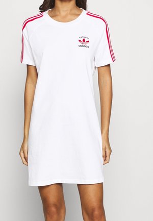 Biała sukienka Adidas z krótkim rękawem, czerwonymi paskami na ramionach i małym logo Adidas England na lewej piersi, noszona przez stojącą osobę.
