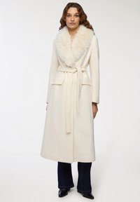 Lungo cappotto crema con colletto in pelliccia sintetica, vita cinturata, tasche anteriori e silhouette dritta. Texture morbida e design elegante.