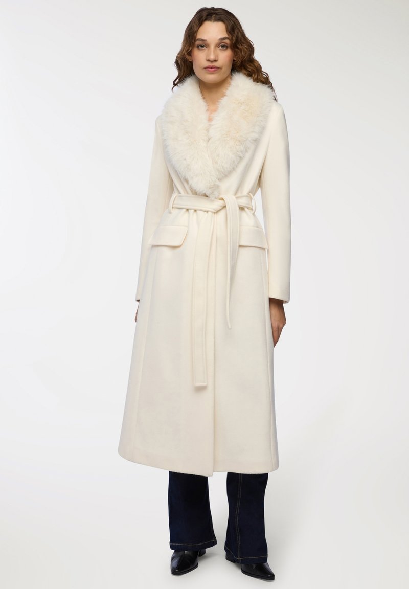 Lungo cappotto crema con colletto in pelliccia sintetica, vita cinturata, tasche anteriori e silhouette dritta. Texture morbida e design elegante.